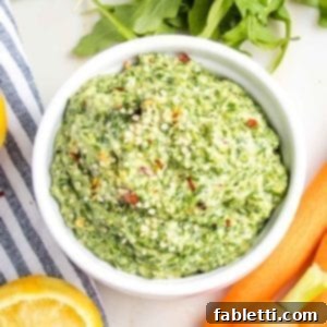 Vibrant Carrot Top Pesto 11 Close up of carrot top pesto in a small white bowl, sprinkled with hemp seeds and crushed red pepper. You can see glimpses of carrots, arugula and lemons surrounding the bowl.