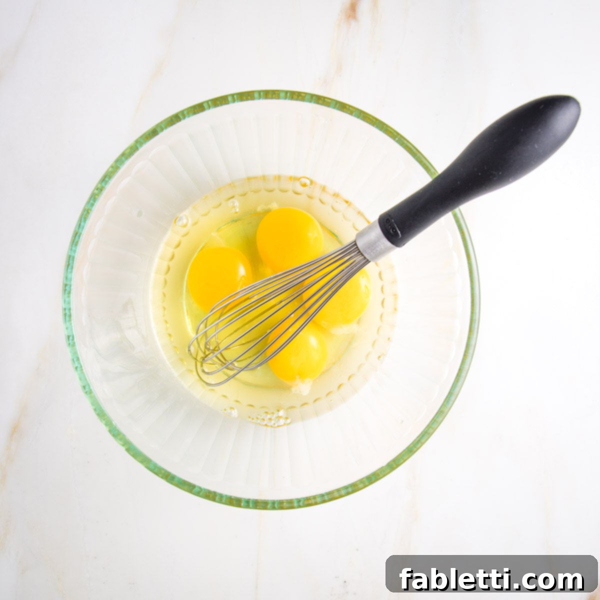 Whole cracked eggs in a glass bowl with a whisk.