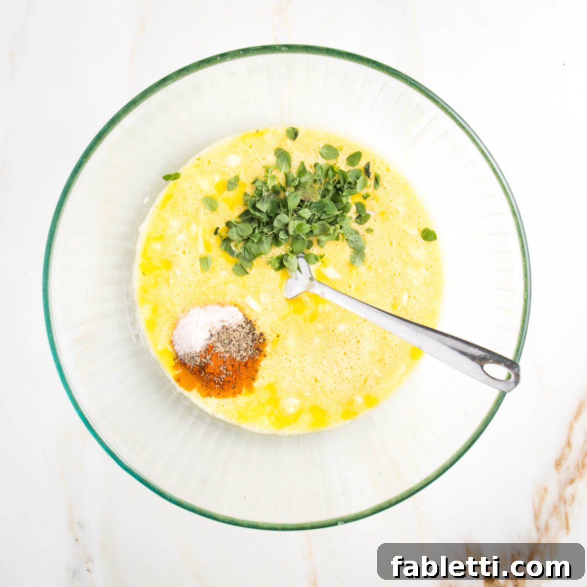 Whisked yellow eggs in a glass bowl with fresh oregano and spices.