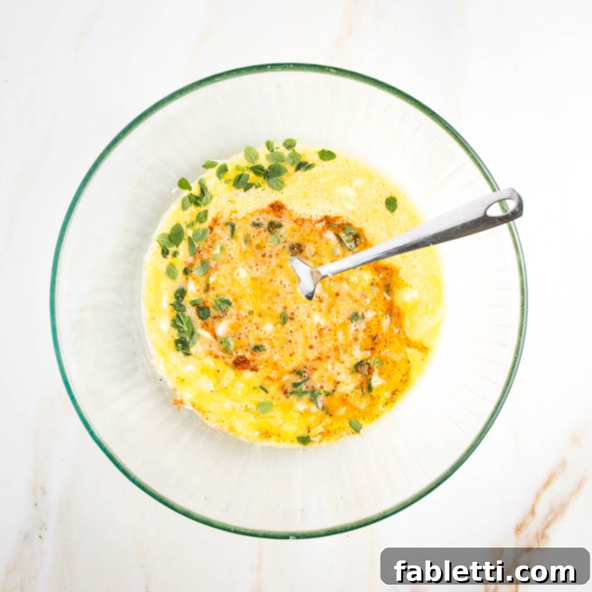 Bowl of bright yellow whisked eggs with paprika and fresh oregano mixing in.