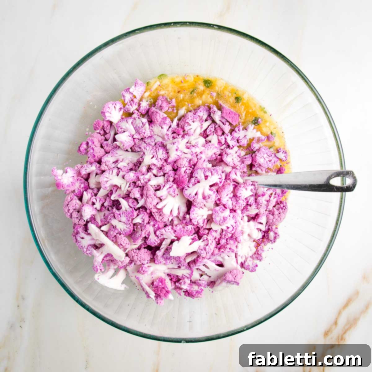 Whisked eggs and spices in a glass bowl, with small pieces of purple cauliflower on top