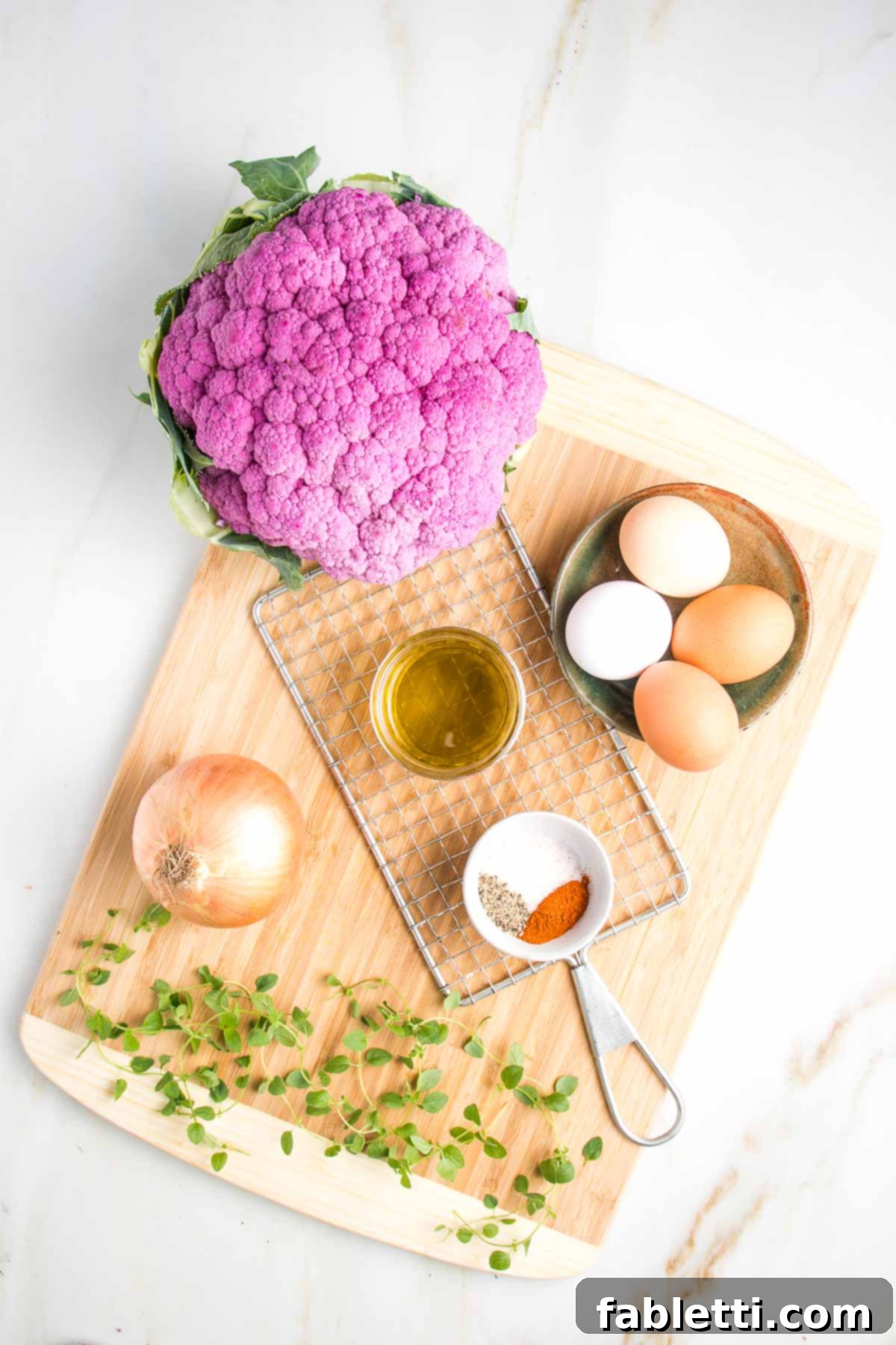 Cauliflower kugel recipe ingredients on a cutting board: large head purple cauliflower, small ceramic dish with 4 eggs of varying colors from white to brown, sprigs of fresh oregano, small white dish with spices, small glass bowl of olive oil, a yellow onion and a potato shredder.