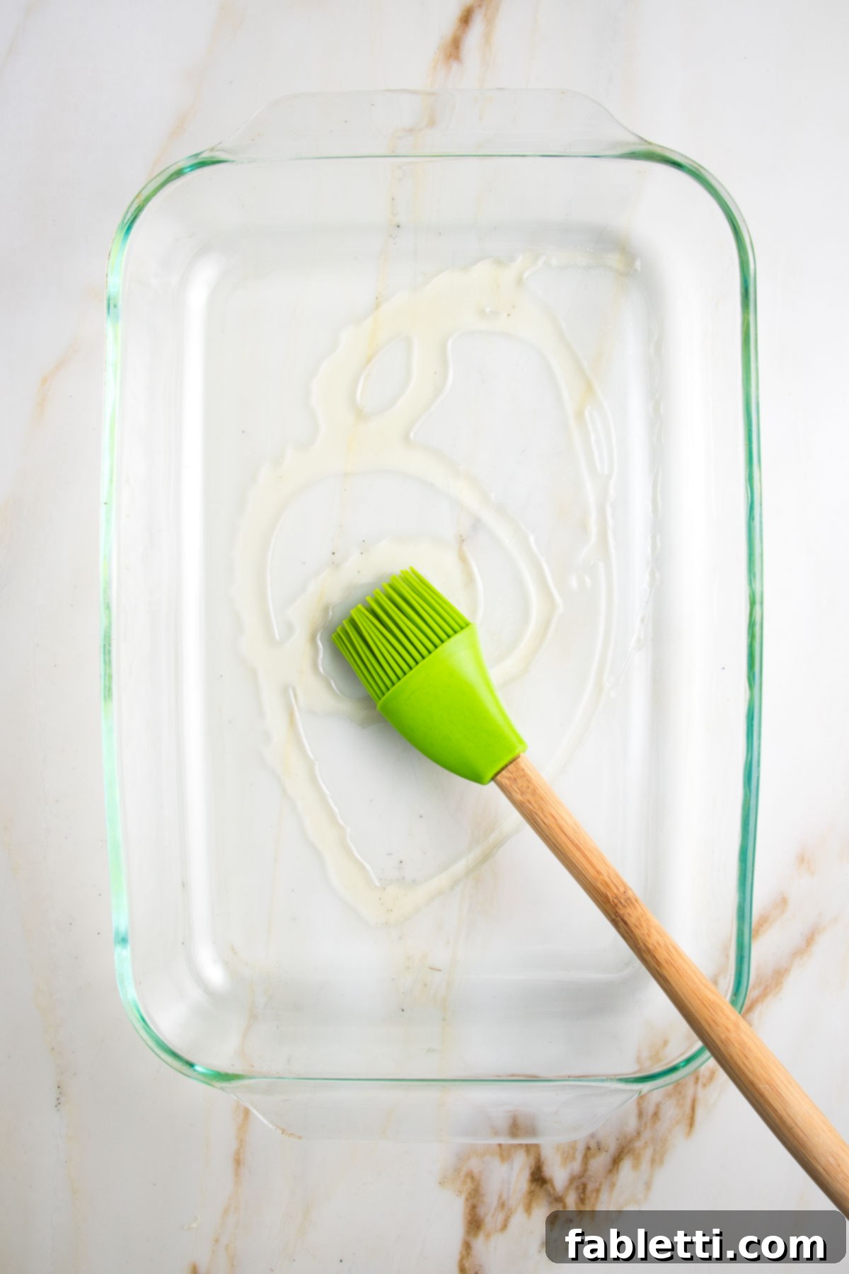 Glass rectangular baking dish swirled with olive oil and a green silicone brush spreading oil.