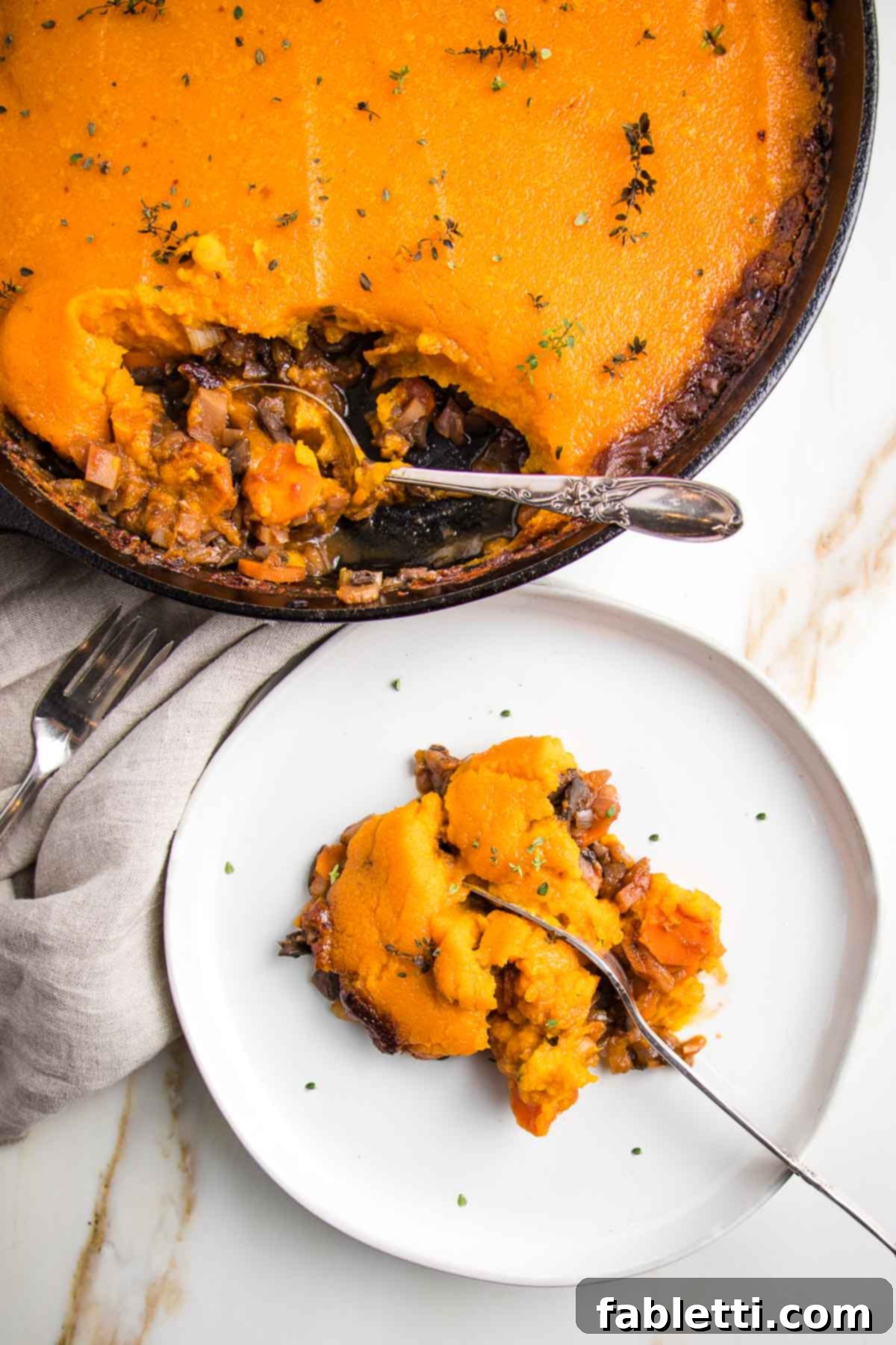 Sun-Kissed Vegan Shepherd's Pie 2 Cast iron skillet with cooked veggie shepherd's pie topped with sweet potato mash. A white rimmed plate is filled with a piece of shepherd's pie and a fork cutting through the middle. of it.