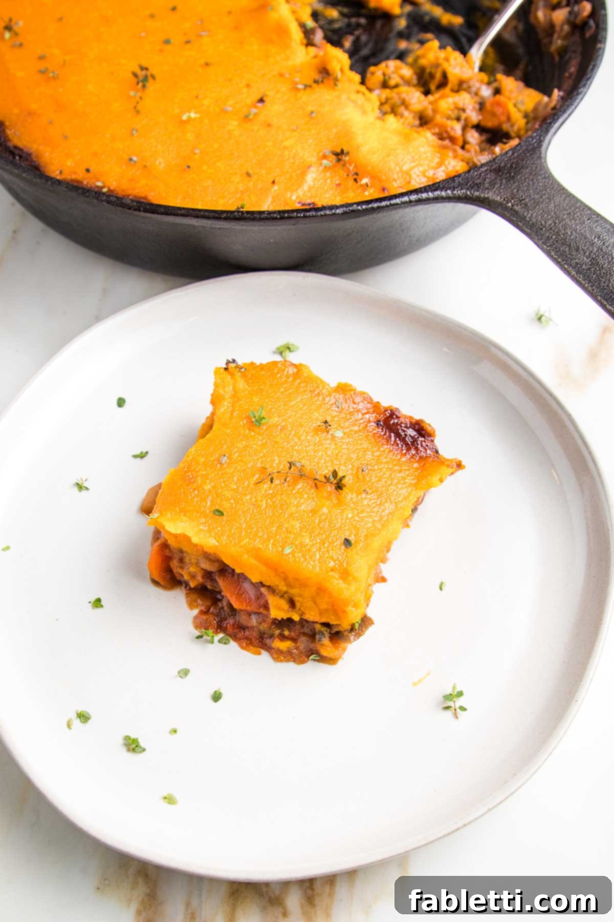 Sun-Kissed Vegan Shepherd's Pie 19 white rimmed plate with piece of shepherd's pie, topped with sweet potato mash. There's a skillet with veggie shepherd's pie in the background.