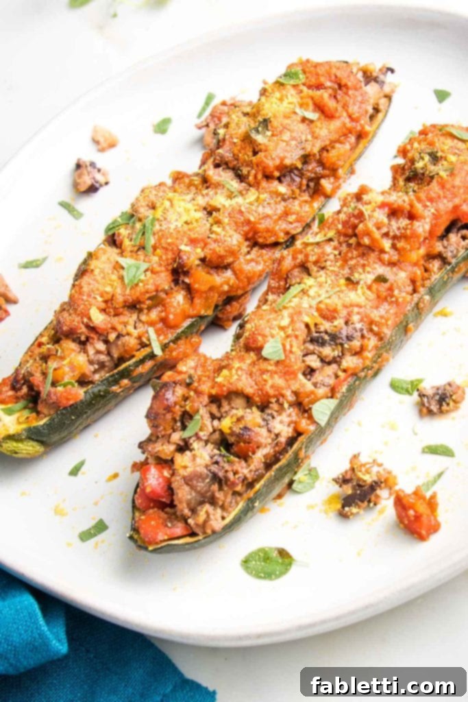 Satisfying Plant-Powered Zucchini Boats 2 Two generously stuffed zucchini boats arranged on a white plate. The plant-based filling mimics ground beef, generously covered with a rich tomato sauce and garnished with fresh herbs and a sprinkle of nutritional yeast. Appetizing crumbs surround the boats, inviting diners to indulge.