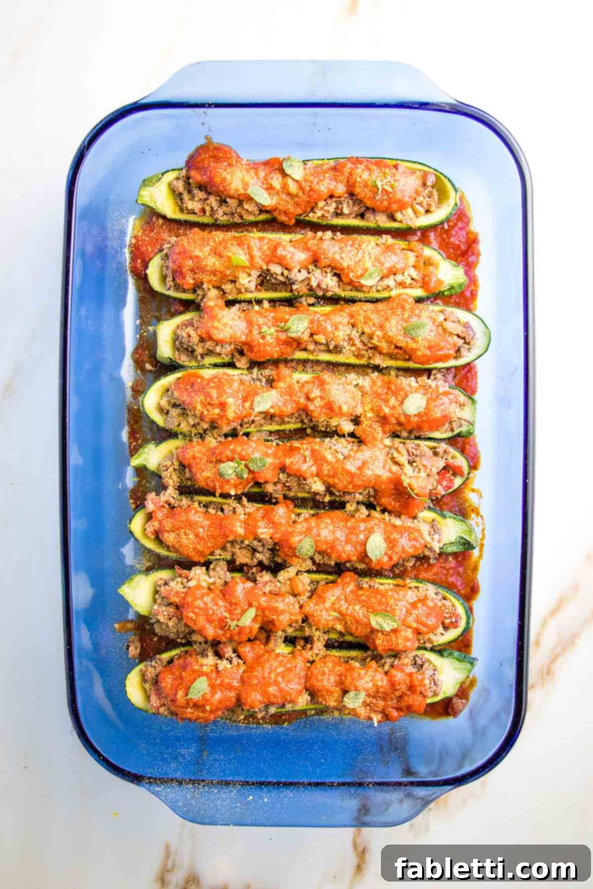 Satisfying Plant-Powered Zucchini Boats 14 A glass baking dish showcasing stuffed zucchini boats, generously covered in marinara sauce and sprinkled with a light dusting of nutritional yeast, ready to be baked to perfection.
