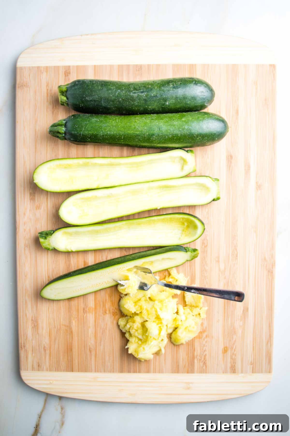 Satisfying Plant-Powered Zucchini Boats 4 Four fresh zucchinis on a cutting board. Two are whole, while two others are halved lengthwise with their insides expertly scooped out. A grapefruit spoon is visible, efficiently removing the pulp from one zucchini half, adding to a pile of scooped-out pulp set aside for the filling.