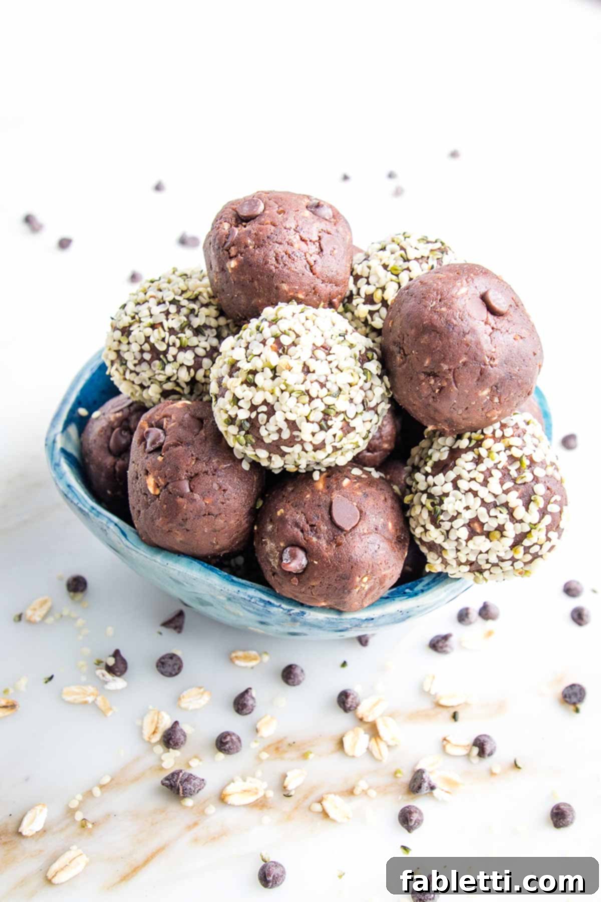 Irresistible Cookie Dough Poppers 2 Small blue bowl filled with chocolate cookie dough bites, some have a hemp seed coating on the outside. The bowl is on a white marble countertop with mini chips sprinkled around.