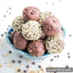 Irresistible Cookie Dough Poppers 16 Small blue bowl filled with chocolate cookie dough balls. Some have a coating of hemp seeds on the outside. There are mini chocolate chips and bits of rolled oats scattered around the bowl on a white marble countertop.