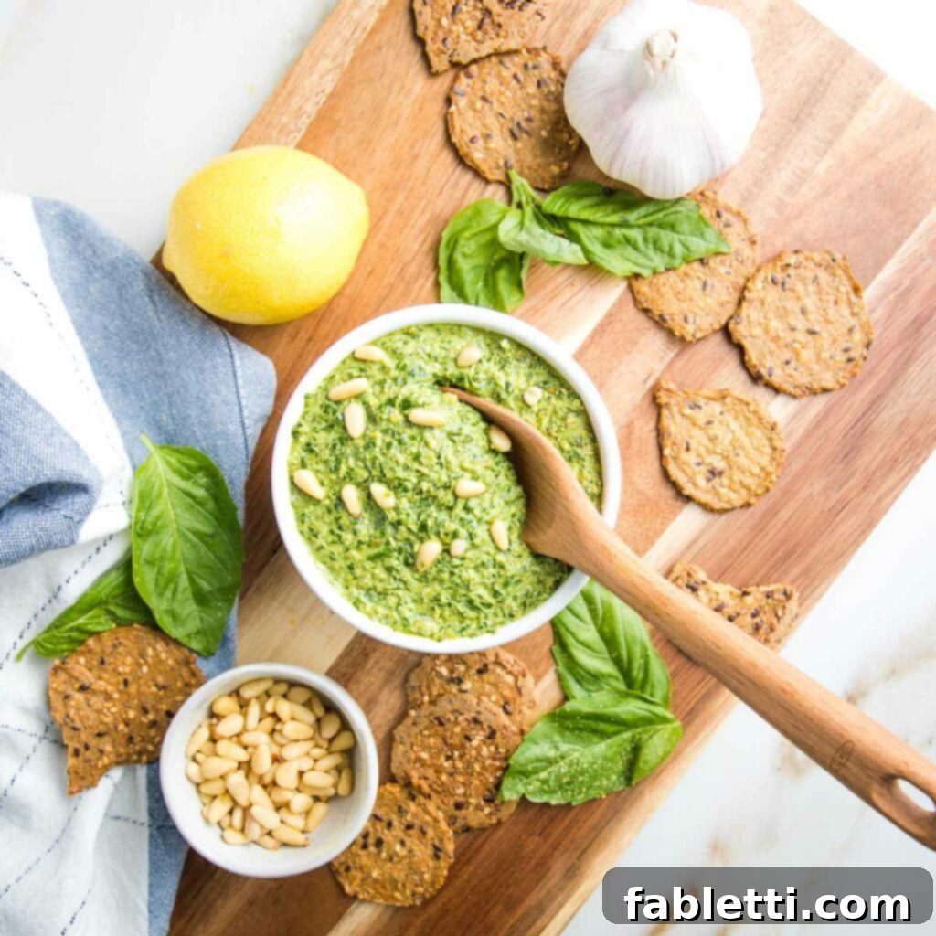 pesto in a small white bowl, on a wooden board. Also shown is a lemon, a head of garlic, some fresh basil, a bowl of pine nuts and some crackers.  Plus a dish towel off to the left. 