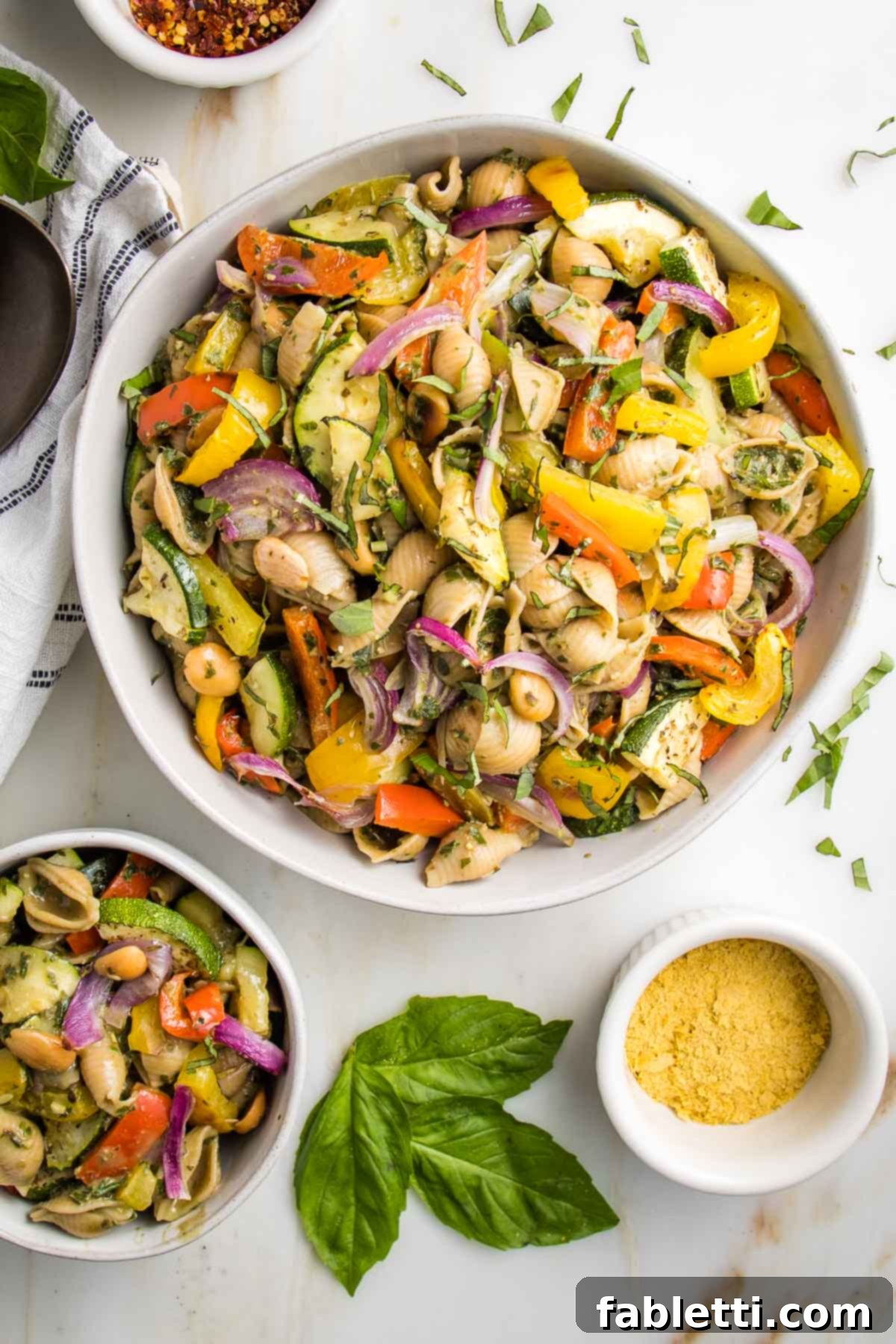 Large white bowl filled with pesto pasta salad with colorful roasted vegetables. There's a small bowl of the pasta on the side along with some fresh basil leaves and a small bowl of nutritional yeast for sprinkling.