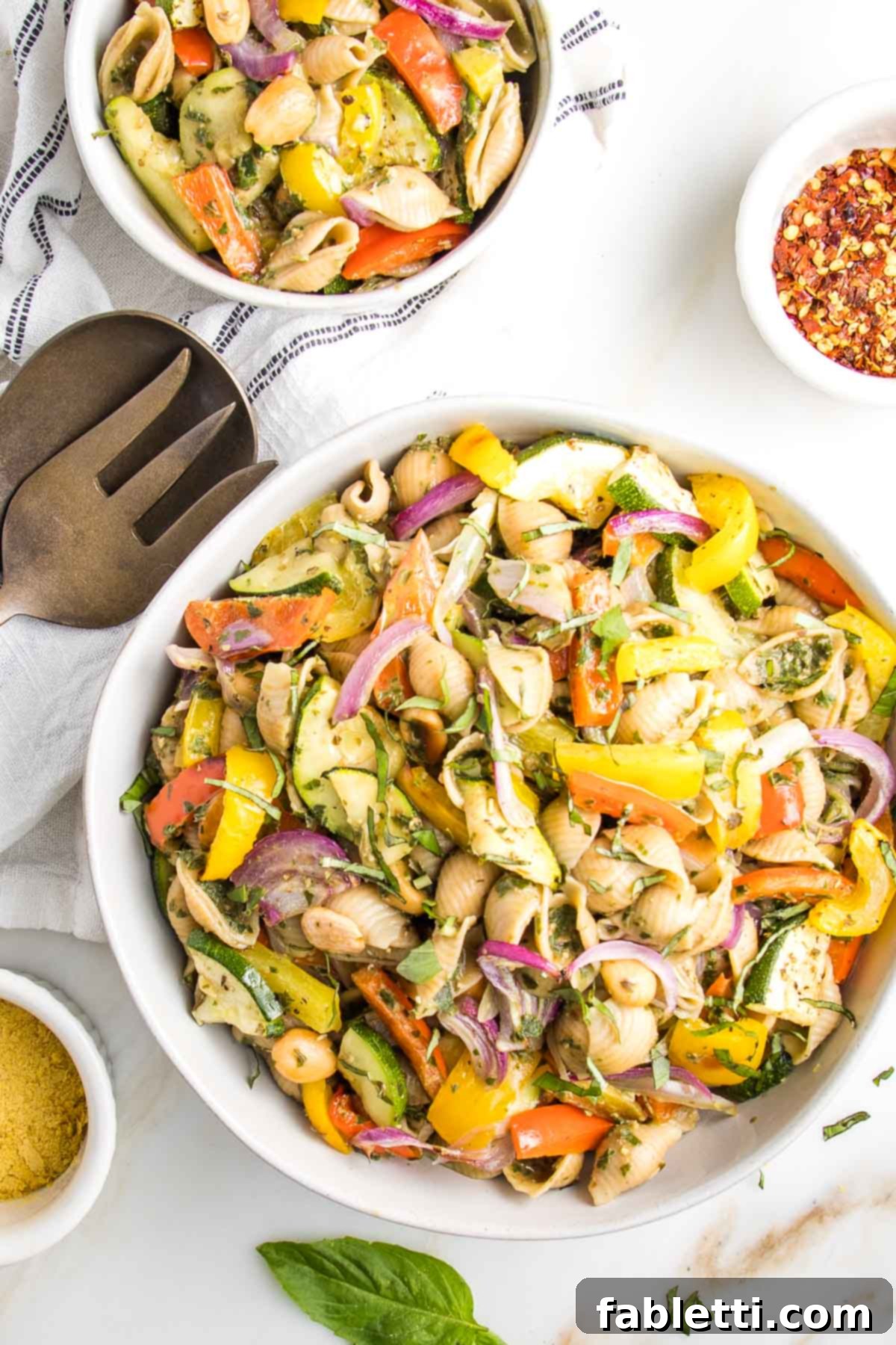 Large white bowl of colorful roasted vegetable pasta salad with pesto sauce. There is also a small bowl of same, some fresh basil leaves, a dish of nutritional yeast and another of crushed red pepper, and a serving fork/spoon.