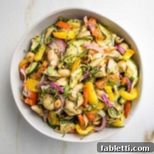 White bowl filled with pasta salad loaded with colorful vegetables and small shells with pesto sauce and fresh slivers of basil.