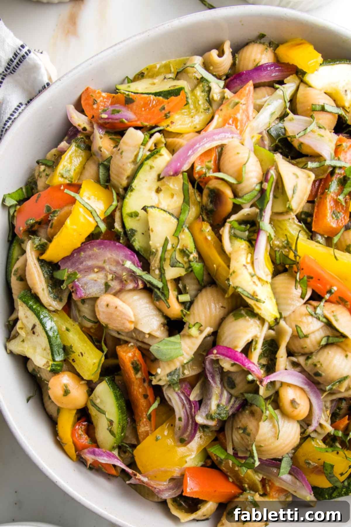 Close up of a bowl of pesto pasta salad with roasted vegetables. It's colorful....red and yellow peppers, purple onions, green zucchini. The pasta is shells, the sauce is green. All in a white bowl.