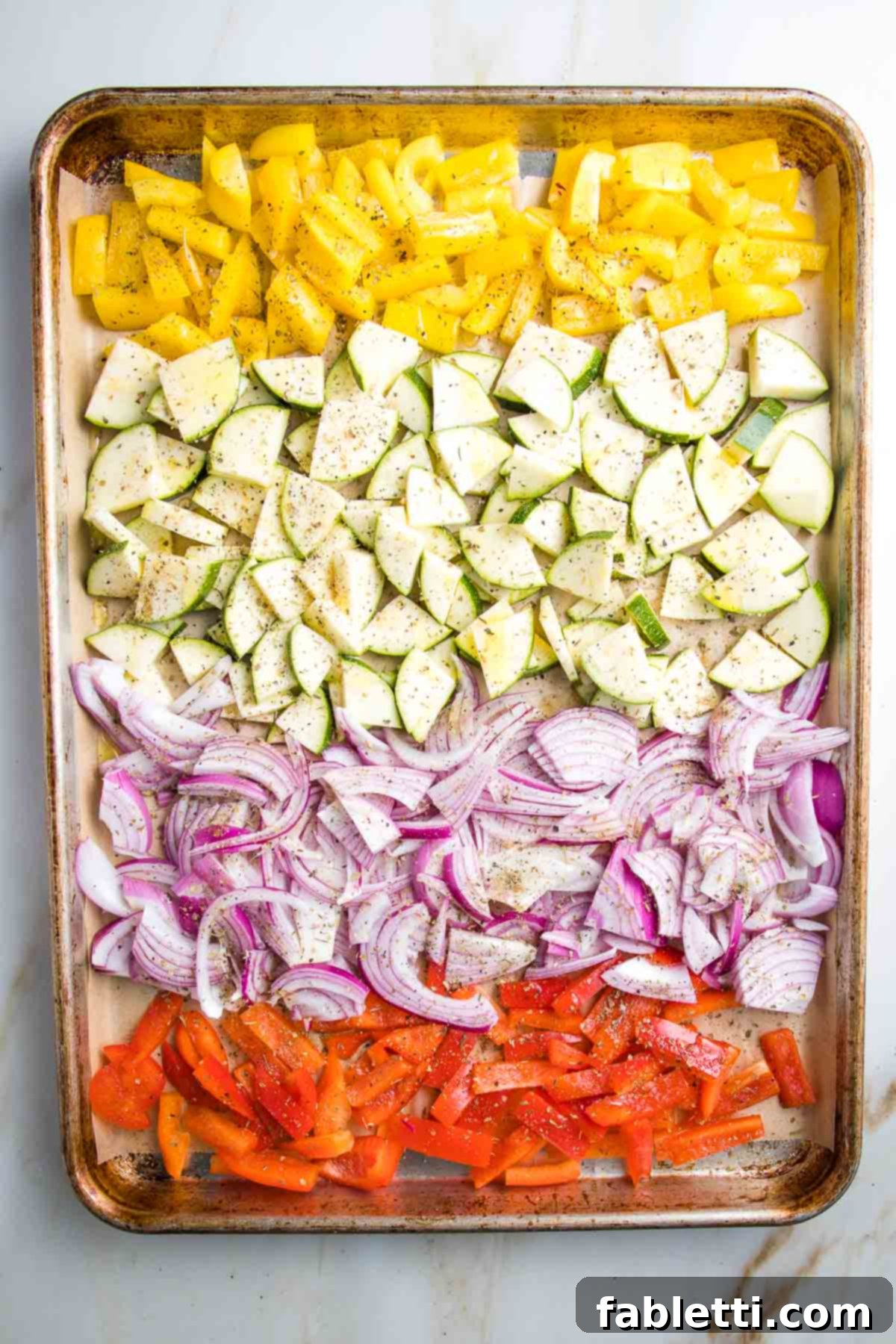 Large rimmed baking tray, lined with parchment paper and loaded with rows of colorful chopped veggies. Yellow peppers, zucchini, purple onions, red peppers.