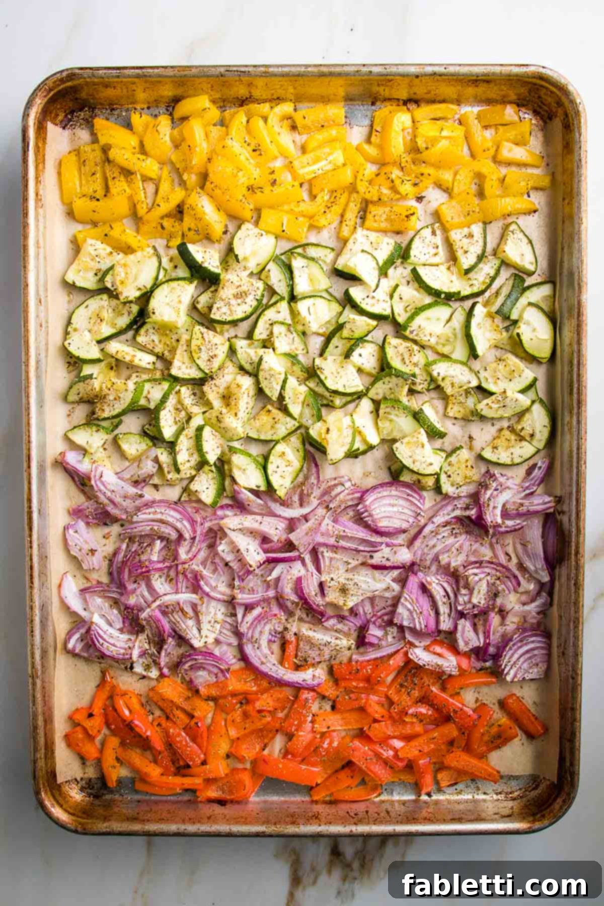 Tray of roasted veggies, sprinkled with italian seasonings. A row each of chopped yellow peppers, then zucchini, purple onions and red peppers.