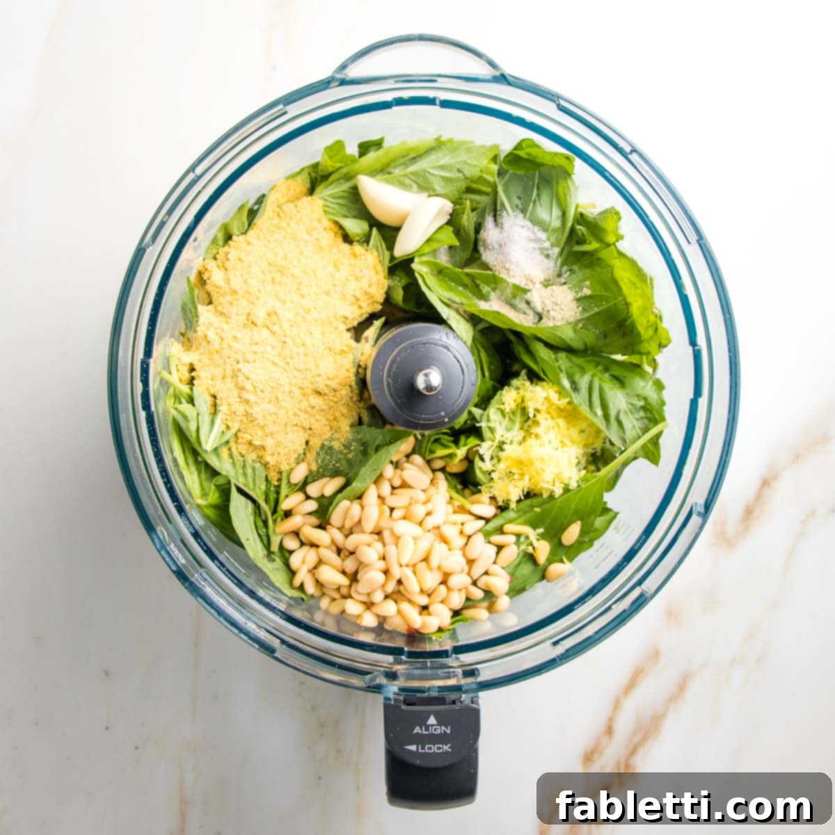 Ingredients for pesto in the bowl of a food processor. Basil, nutritional yeast, pine nuts, lemon zest and juice, garlic.