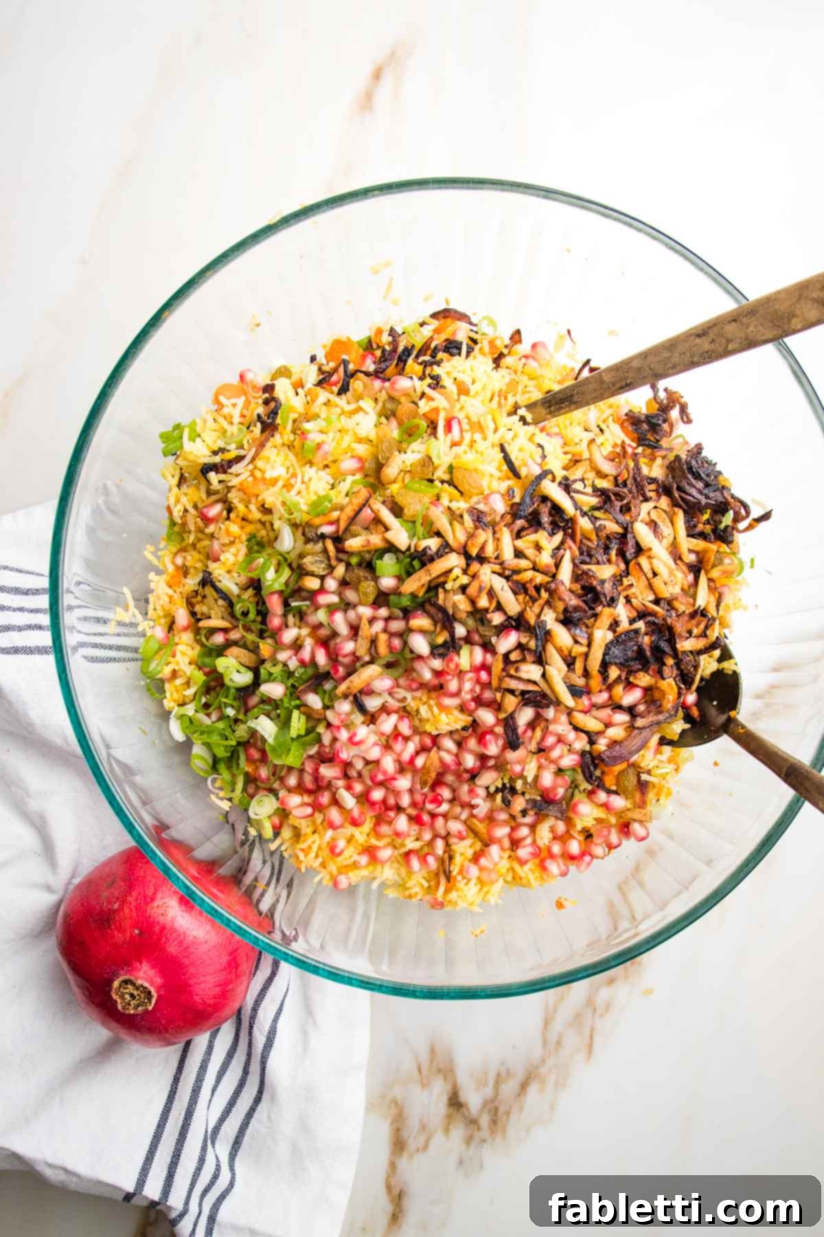 Large glass bowl filled with yellow rice, sliced scallions, pomegranate seeds, golden raisins and caramelized onions.