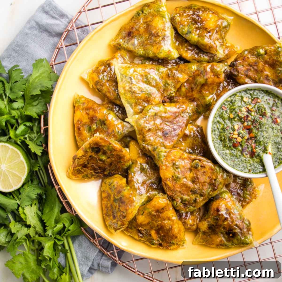 Crispy Baked Samosas with Zesty Cilantro Chutney 17 Baked samosas in small triangles on a yellow dish with a small white bowl filled with green chutney and a bunch of cilantro on the side.