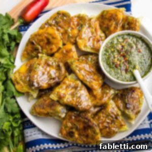 Crispy Baked Samosas with Zesty Cilantro Chutney 19 White rimmed round plate filled with appetizer sized baked samosas plus a small white dish filled with green chutney. The plate is on a blue patterned napkin and is surrounded by a bunch of cilantro and a red chili pepper