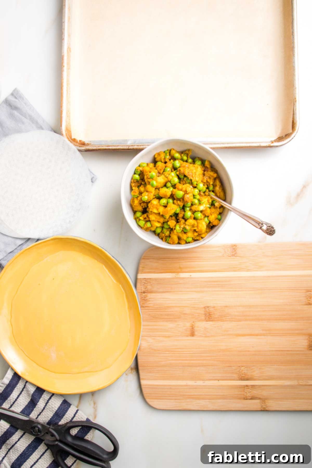 Crispy Baked Samosas with Zesty Cilantro Chutney 9 Everything ready to begin making samosas: rimmed baking tray lined with parchment, stack of rice paper wrappers, rimmed dish filled with water, samosa filling, small wooden board, kitchen scissors and a dish towel.