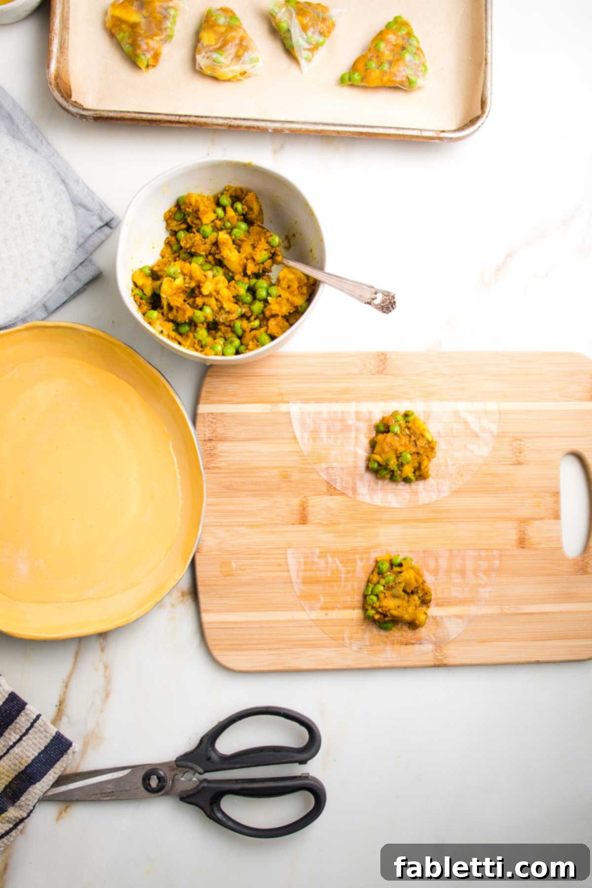 Crispy Baked Samosas with Zesty Cilantro Chutney 10 Rimmed baking sheet lined with parchment, a bowl of potato samosa filling, a yellow dish filled with water, a pair of kitchen scissors, and a wooden board with two half moons of rice paper each with a scoop of filling.