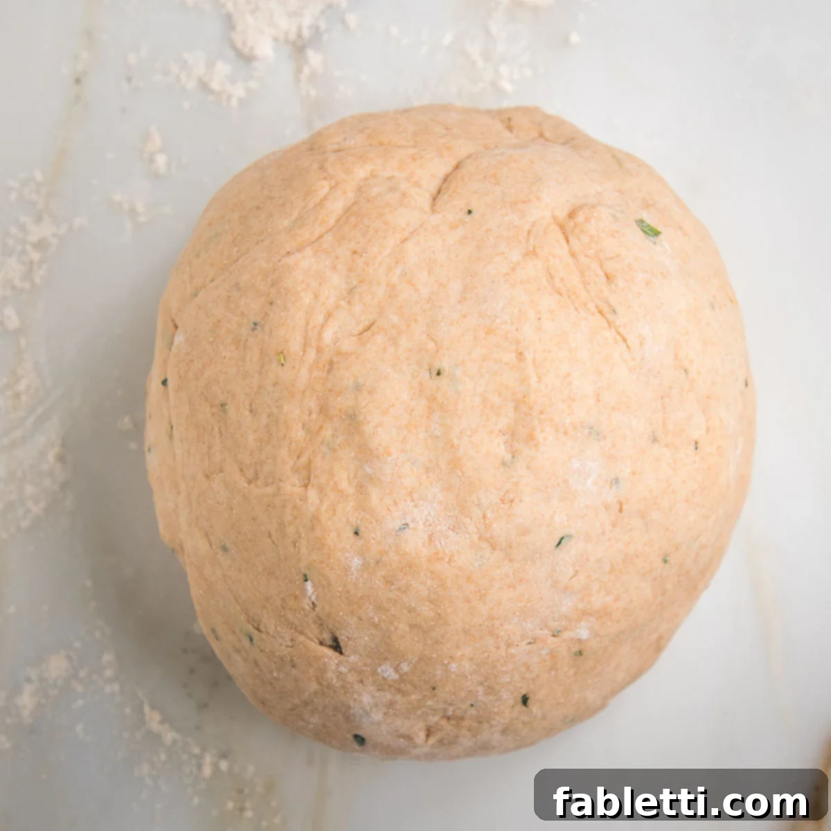 Fluffy Vegan Dinner Rolls 13 dough shaped into a large round ball. You can see flecks of the fresh herbs in the dough and flour sprinkled onto the counter.