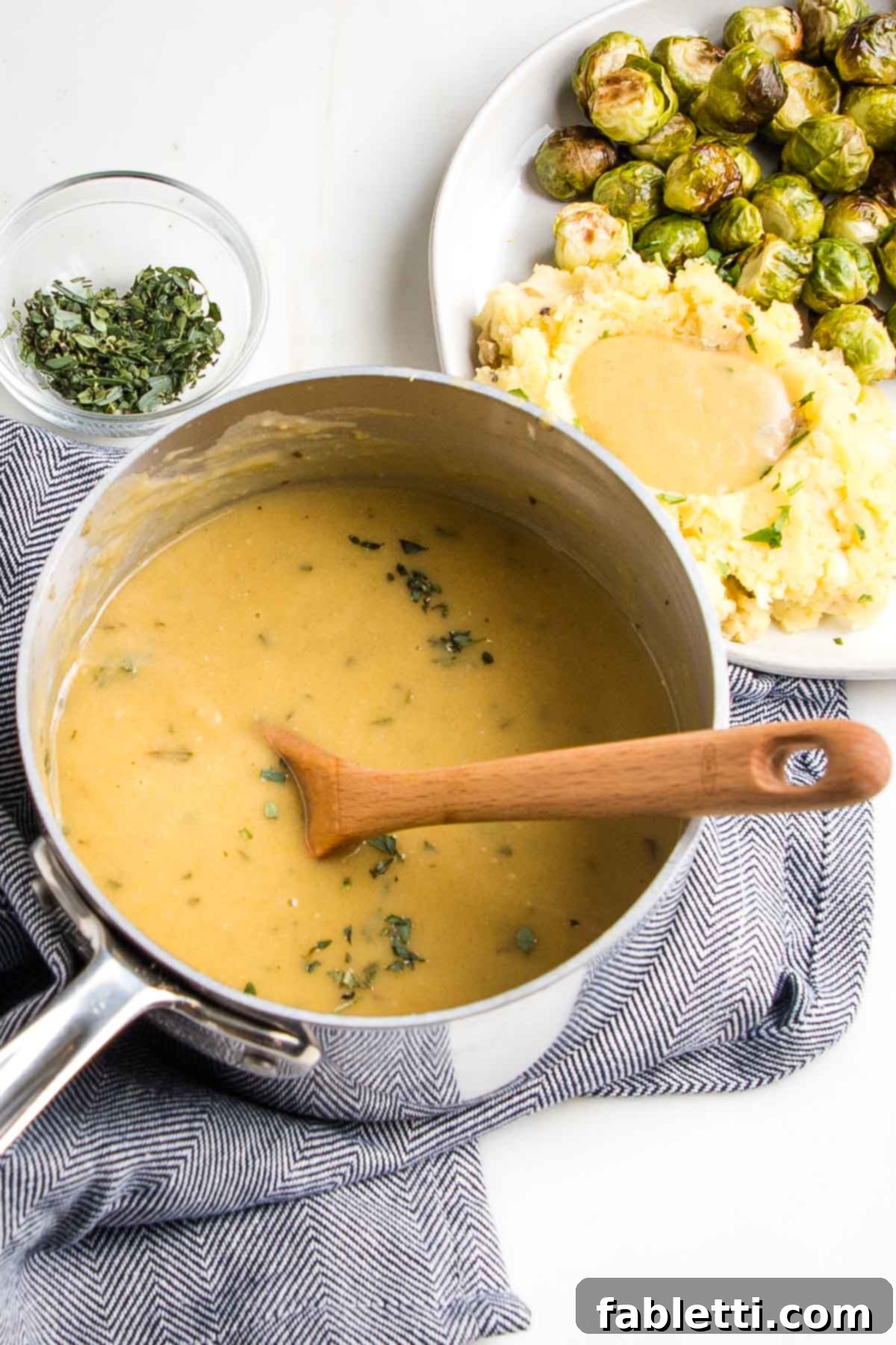 Whip Up Perfect Vegan Gravy 2 A small saucepan brimming with golden vegan gravy, a wooden spoon gently stirring, positioned beside a plate of creamy mashed potatoes generously topped with gravy and vibrant green Brussels sprouts. This image evokes warmth and comfort, highlighting the rich texture and inviting color of the homemade plant-based sauce.