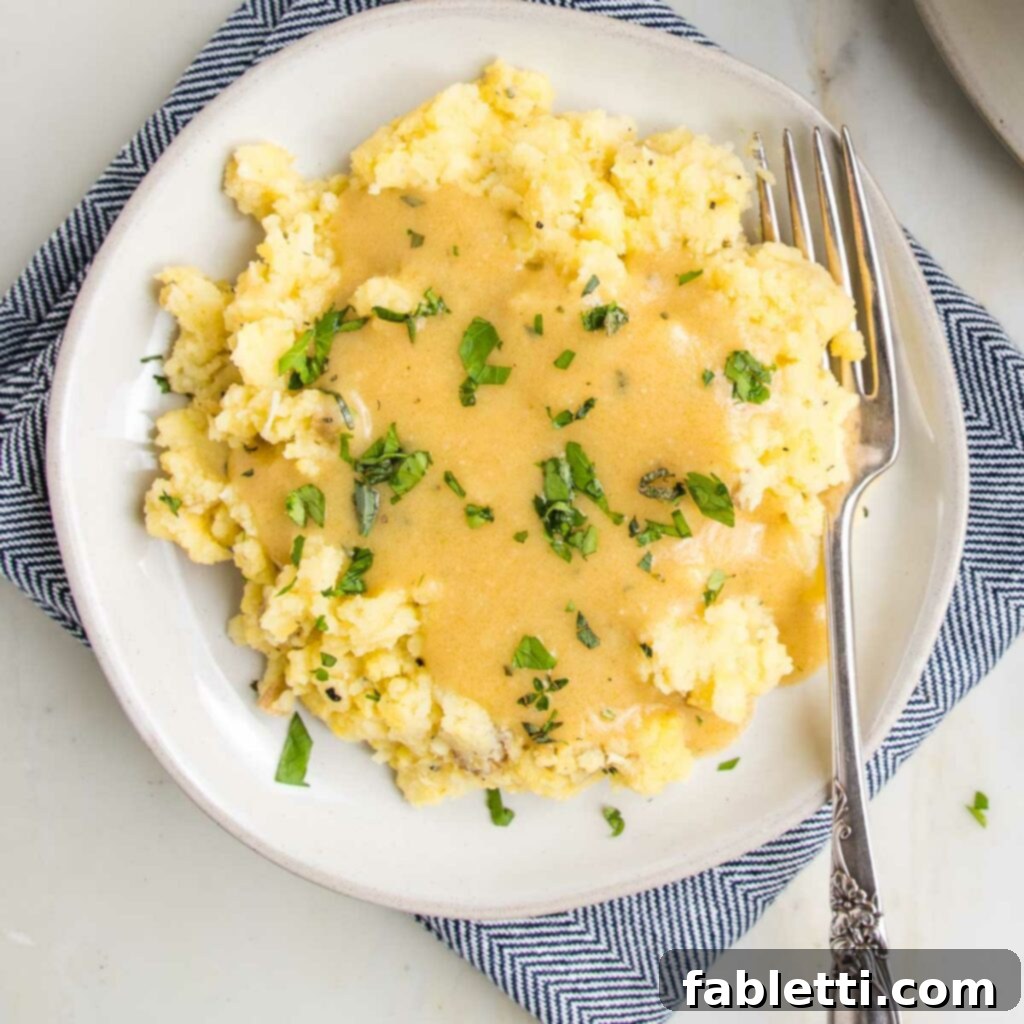 Whip Up Perfect Vegan Gravy 11 A pristine white plate holds a generous mound of fluffy mashed potatoes, lavished with golden vegan gravy and elegantly garnished with a sprinkle of fresh, vibrant green herbs. The lighting highlights the rich texture of the potatoes and the glossy, appetizing sheen of the gravy.