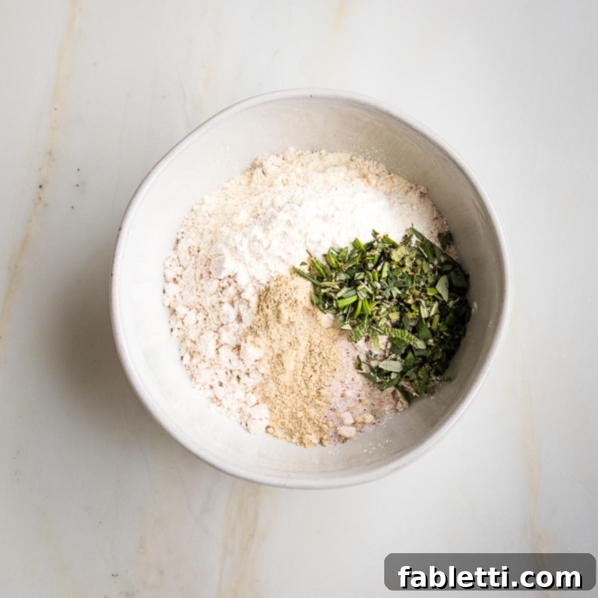 Whip Up Perfect Vegan Gravy 5 A small white bowl filled with a dry mixture of flour, finely chopped fresh herbs, arrowroot powder, salt, white pepper, garlic powder, and onion powder, ready for the next step in making vegan gravy.