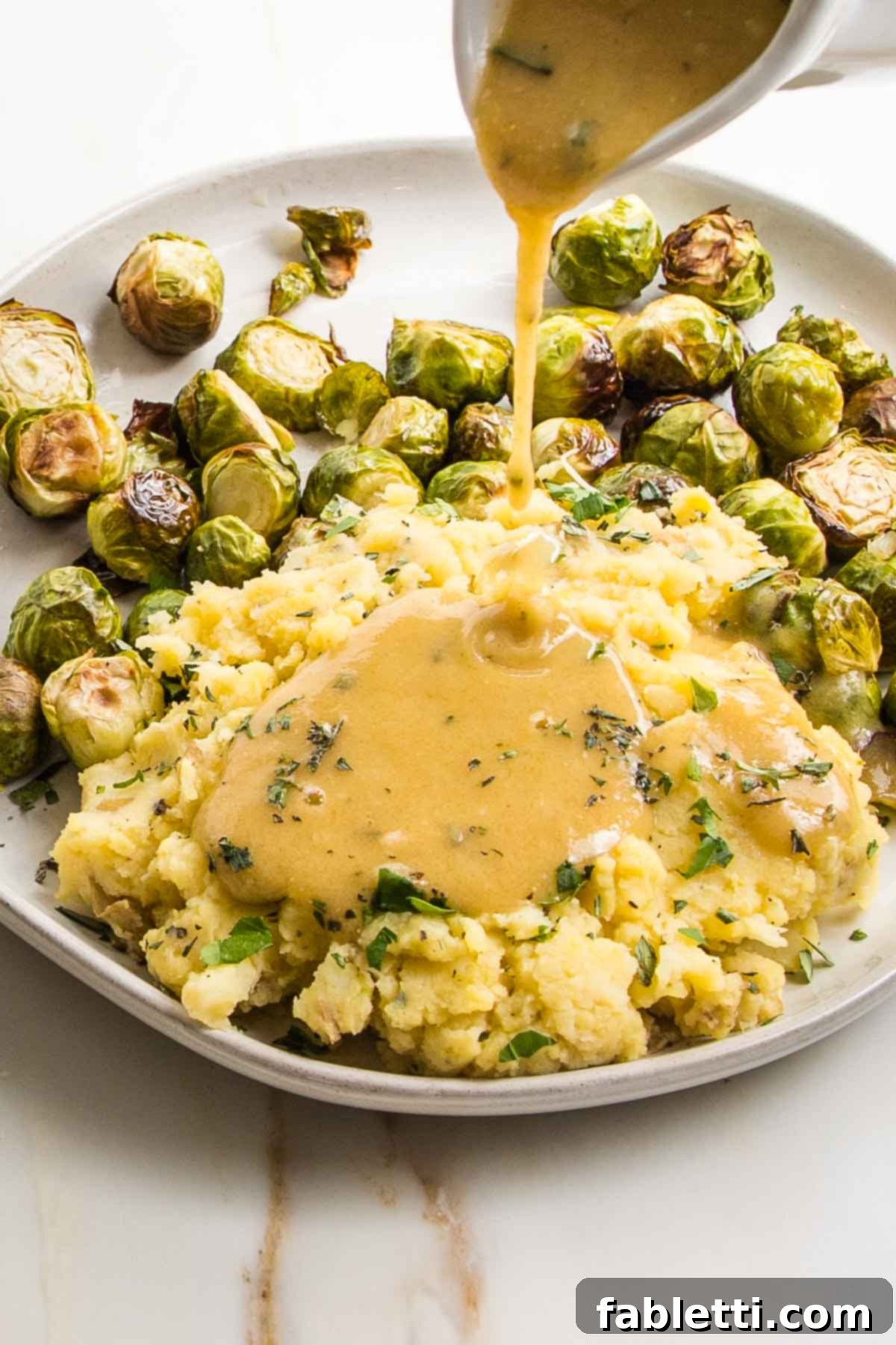Whip Up Perfect Vegan Gravy 10 A stylish white pitcher pouring rich, golden vegan gravy over a generous serving of mashed potatoes and green Brussels sprouts on a white plate. The gravy cascades smoothly, inviting diners to enjoy a hearty, plant-based meal.