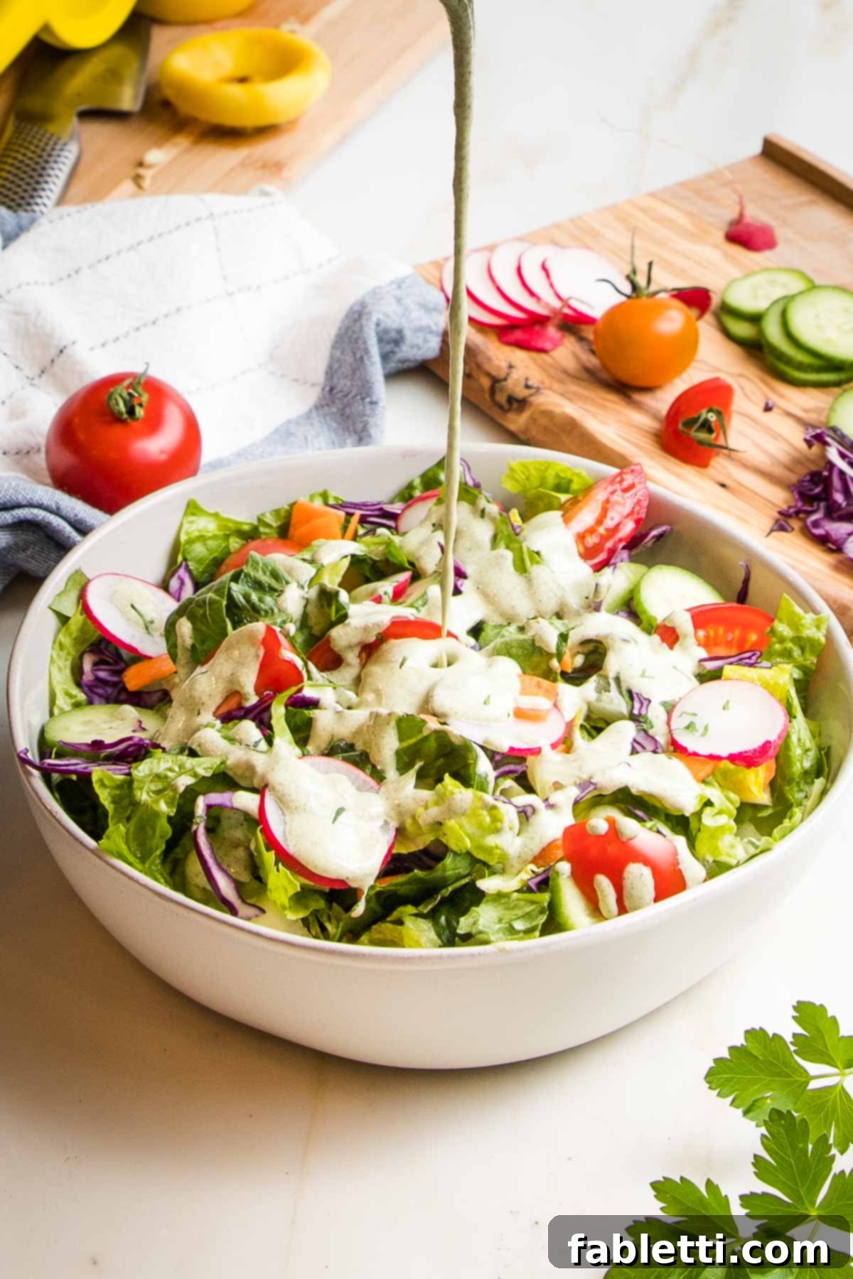 Deliciously Healthy Homemade Ranch 2 A fresh, simple salad featuring crisp romaine lettuce, thinly sliced radishes, vibrant carrots, refreshing cucumbers, and ripe tomatoes in a pristine white bowl. A luscious stream of creamy vegan ranch dressing is being poured generously over the salad, creating inviting puddles in the center.