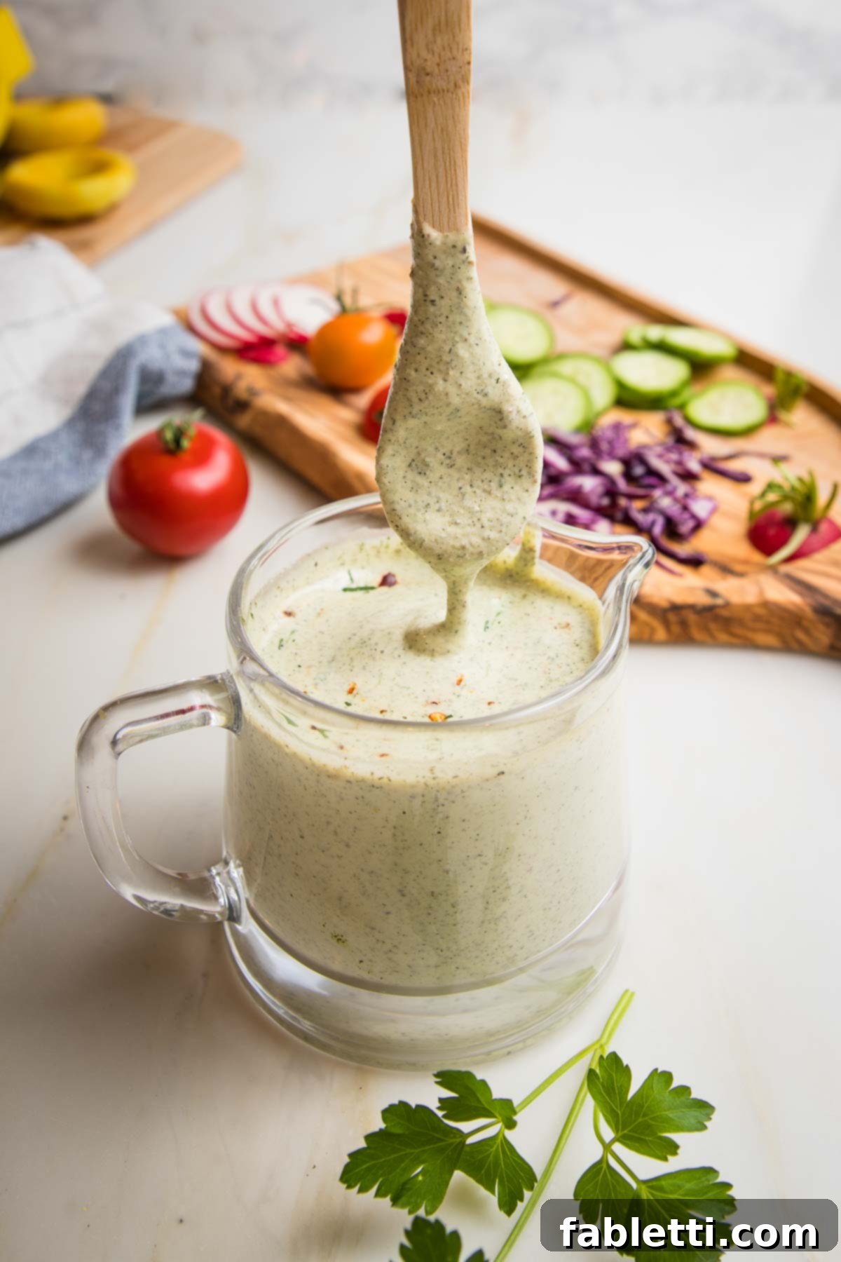 Deliciously Healthy Homemade Ranch 7 A small glass pitcher filled with homemade healthy ranch dressing, with a wooden spoon partially submerged, illustrating its pourable yet creamy consistency. In the soft-focused background, an assortment of colorful fresh vegetables are casually arranged on a wooden cutting board, hinting at healthy dipping options.