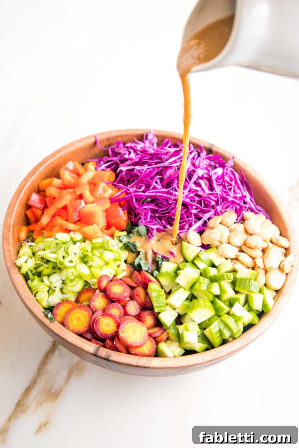 Rustic Tuscan Chopped Kale Medley 11 Wooden Salad bowl with sections of colorful chopped veggies cabbage, cucumbers, carrots, red peppers, scallions and marcona almonds) and a small white pitcher of light brown dressing pouring over the center of the bowl.
