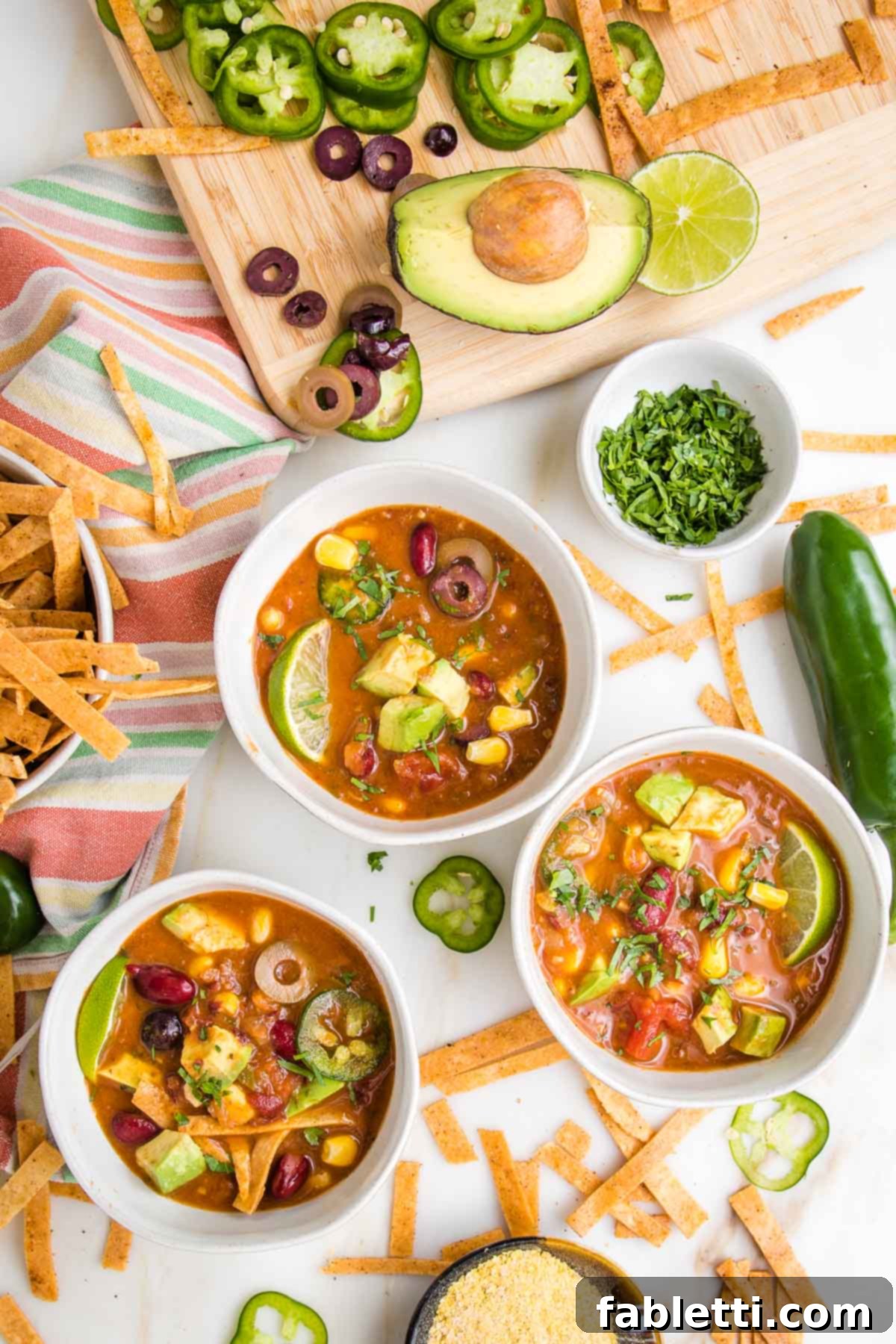 Hearty Vegan Taco Soup with Crispy Tortilla Strips 2 Three bowls of colorful vegan taco soup, with chunks of avocaod, slices of jalapeno, some tortilla strips, a bowl of chopped cilantro, some nutritional yeast and the edge of a cutting board filled with sliced olives, jalapenos and an avocado.