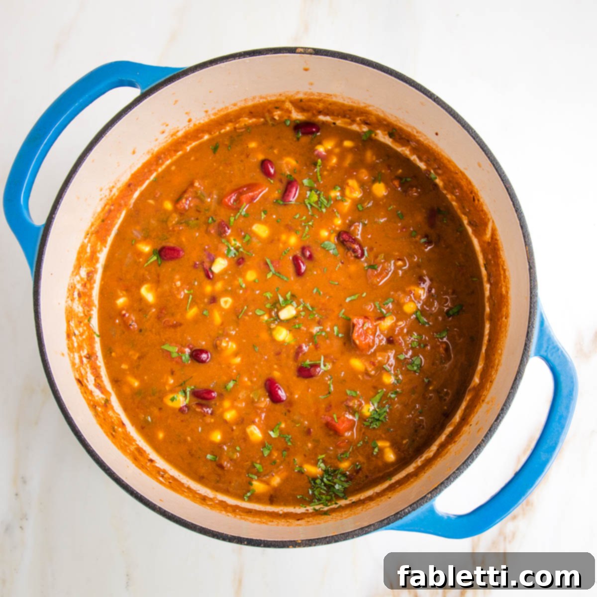 Hearty Vegan Taco Soup with Crispy Tortilla Strips 17 colorful taco soup in a blue dutch oven. Tomato base and you can see red kidney beans, fresh chopped cilantro, corn, pieces of tomatoes and onions.