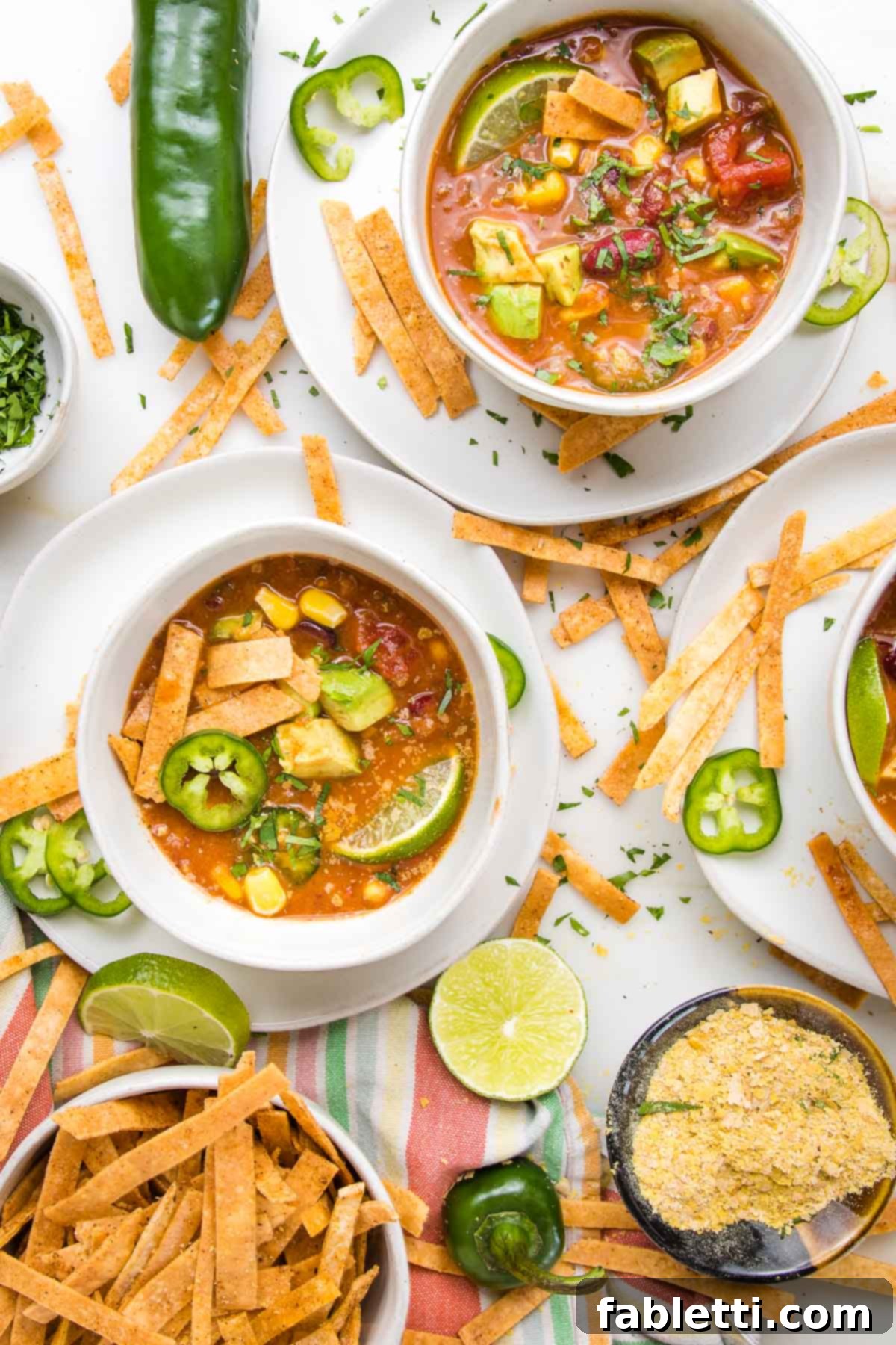 Hearty Vegan Taco Soup with Crispy Tortilla Strips 20 Vegan Taco Soup in white bowls, with toppings...sliced jalapenos, tortilla strips, chunks of avocado, fresh lime wedge. There is also a bowl of tortilla strips, a small dish of nutritional yeast and an active scene of taco soup chaos.