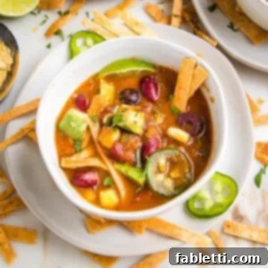 Hearty Vegan Taco Soup with Crispy Tortilla Strips 21 White bowl with plate underneath filled with vegan taco soup. It's colorful, with yellow corn, green jalapenos, chunks of avocado, red kidney beans and crispy tortilla strips. You also can see additional toppings strewn about on the white counter.