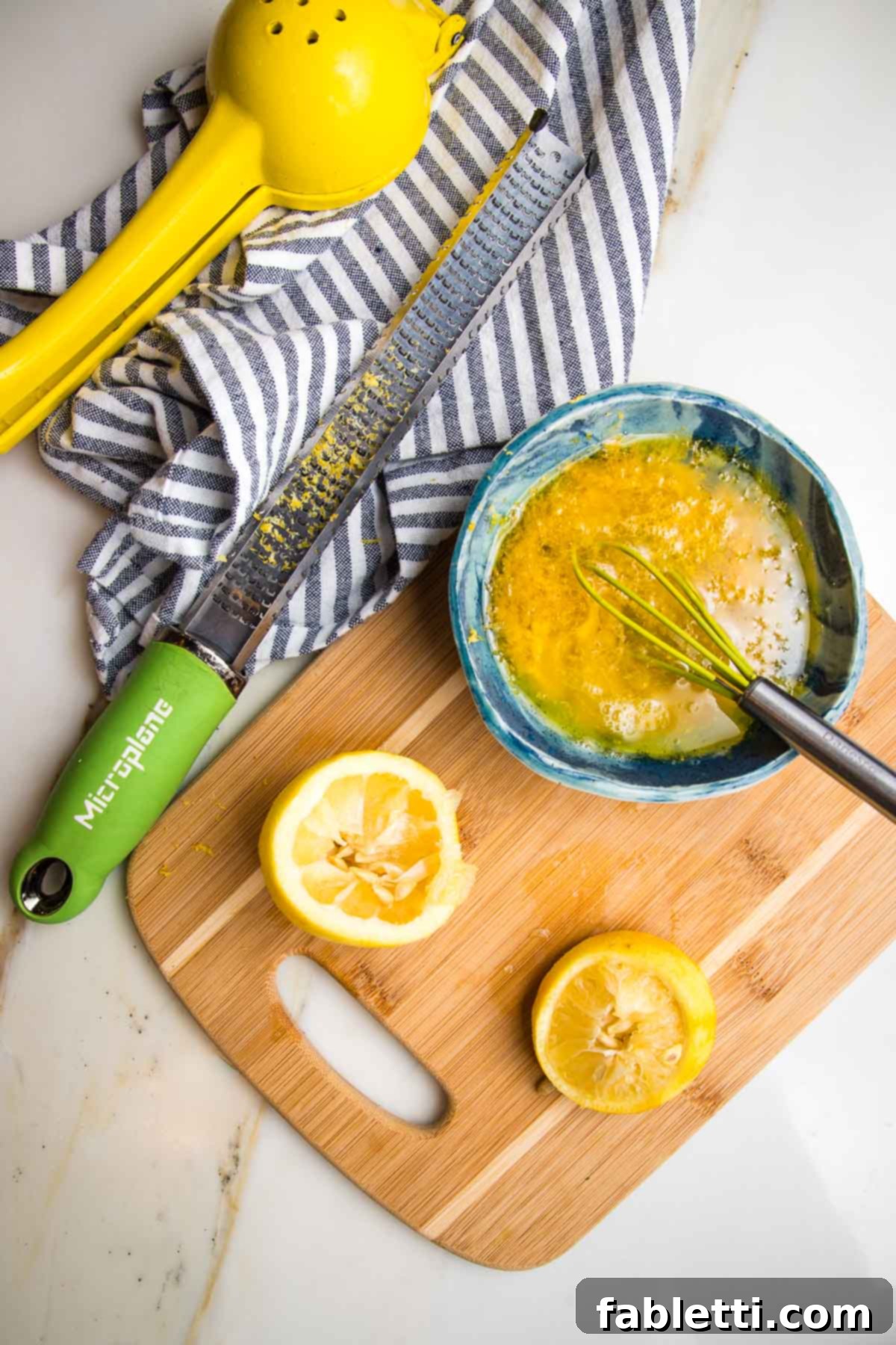 Zesty Kidney Bean Salad 7 oily dressing whisked together in teal bowl. Two halves of a lemon already zested and squeezed lie on a cutting board. A zester with lemon zest on it on a blue striped towel.