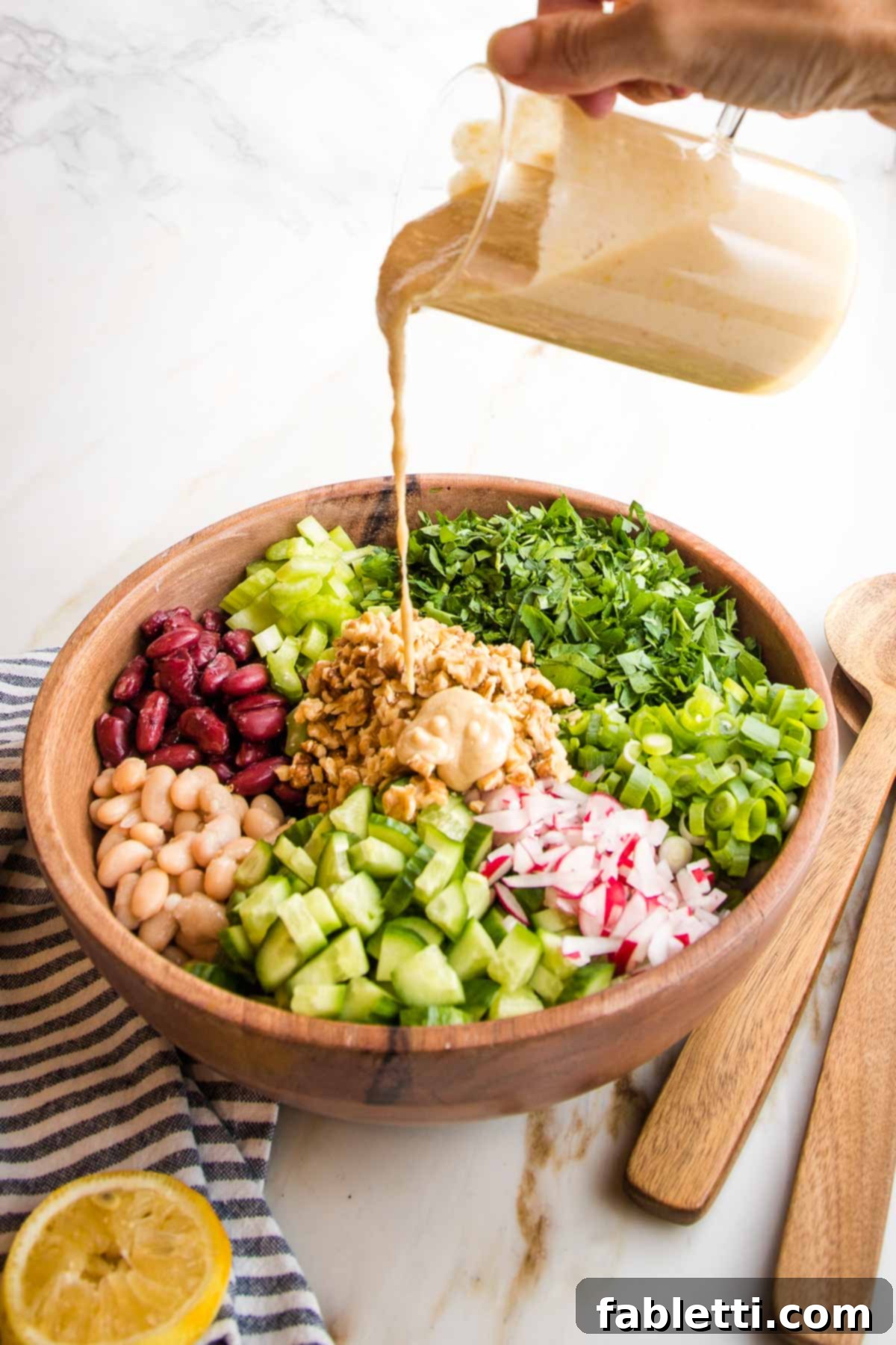 Zesty Kidney Bean Salad 9 Wooden bowl with vibrant chopped vegetable sections. Lemon tahini dressing is being poured over the bowl at the center where the walnuts are.