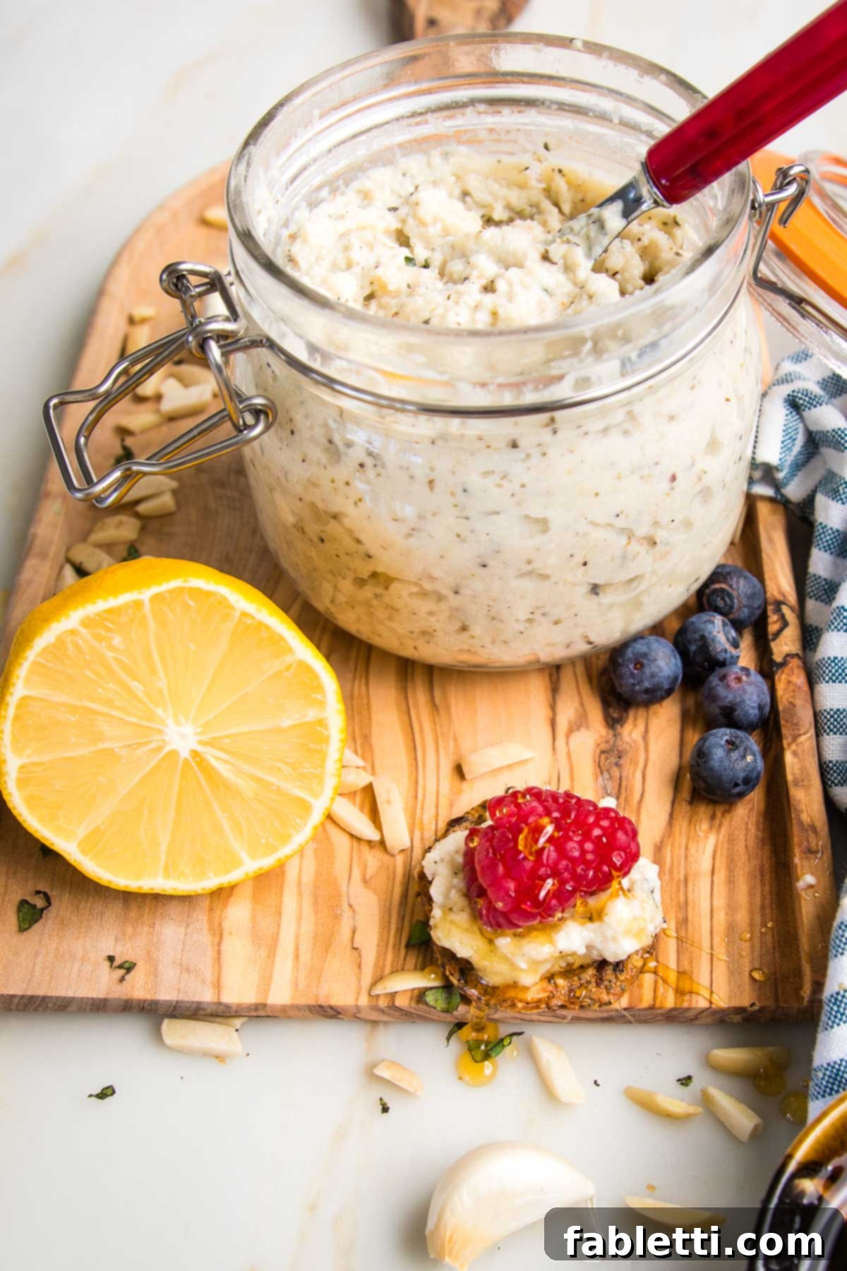 Jar of vegan ricotta and a spreader. A cracker spread with ricotta and a fresh raspberry, drizzled with honey and a few blueberries, plus half a lemon and a few slivered almonds and cloves of garlic.