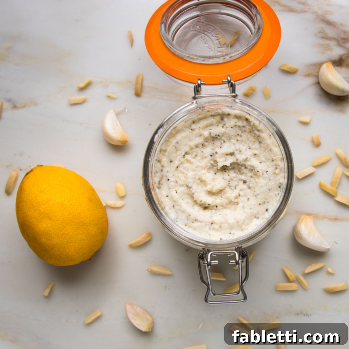 Glass storage jar filled with vegan ricotta cheese. A lemon, slivered almonds and cloves of garlic are scattered on the counter around the cheese jar.