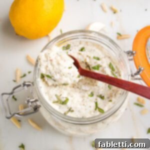 glass jar filled with vegan ricotta, sprinkled with fresh herbs...a spoon is scooping some out. THere are slivered almonds, garlic cloves and a lemon in the background.