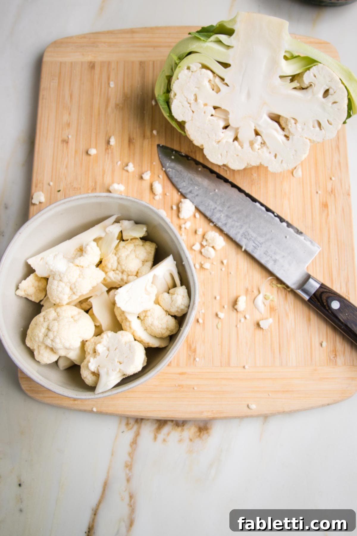 Cauliflower cut in half. One half is left whole in the background and a sharp knife is show that cut the other half into florets that are in a small white bowl. All is on a wooden board.