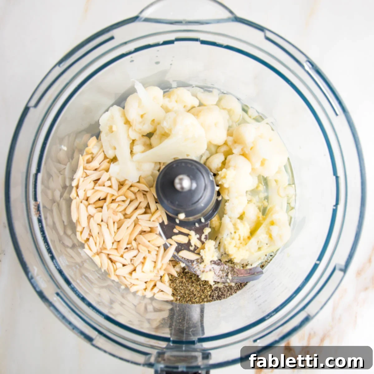 Food processor bowl with slivered almonds, steamed cauliflower, zested garlic, spices, lemon juice, olive oil and water.