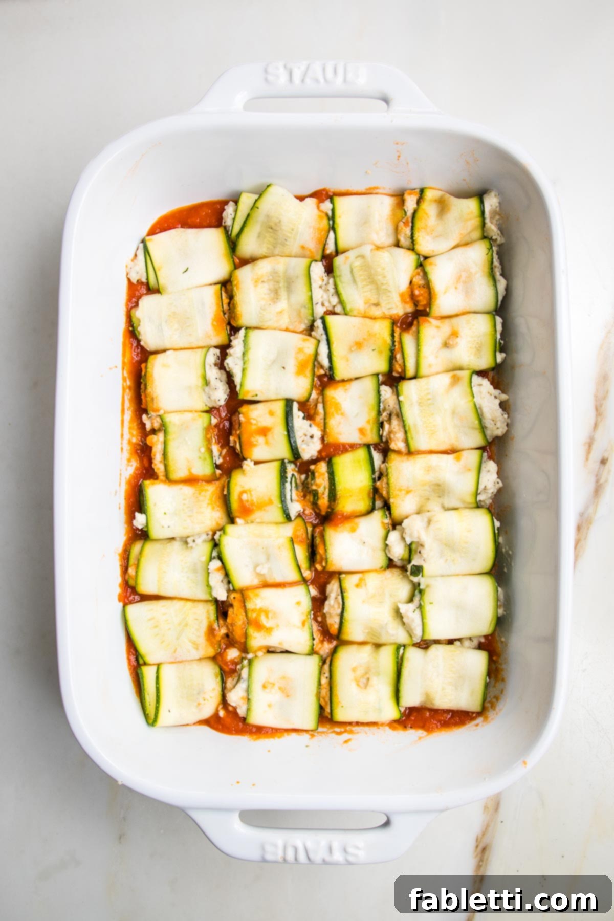 Plant-Based Zucchini Ricotta Rolls 11 White baking pan spread with marinara and topped with zucchini ricotta roll ups.