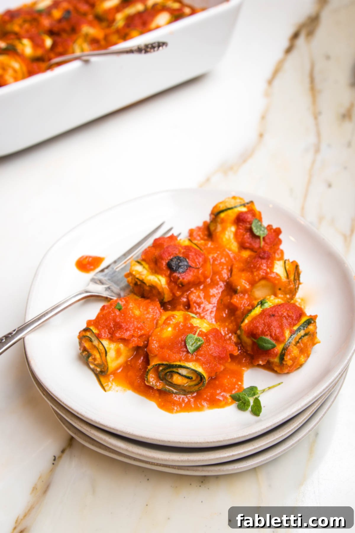 Plant-Based Zucchini Ricotta Rolls 14 White rimmed plate filled with rolled up zucchini lasagna rolls covered in marinara sauce. Fork on the plate. Casserole dish filled with more visible in the background.