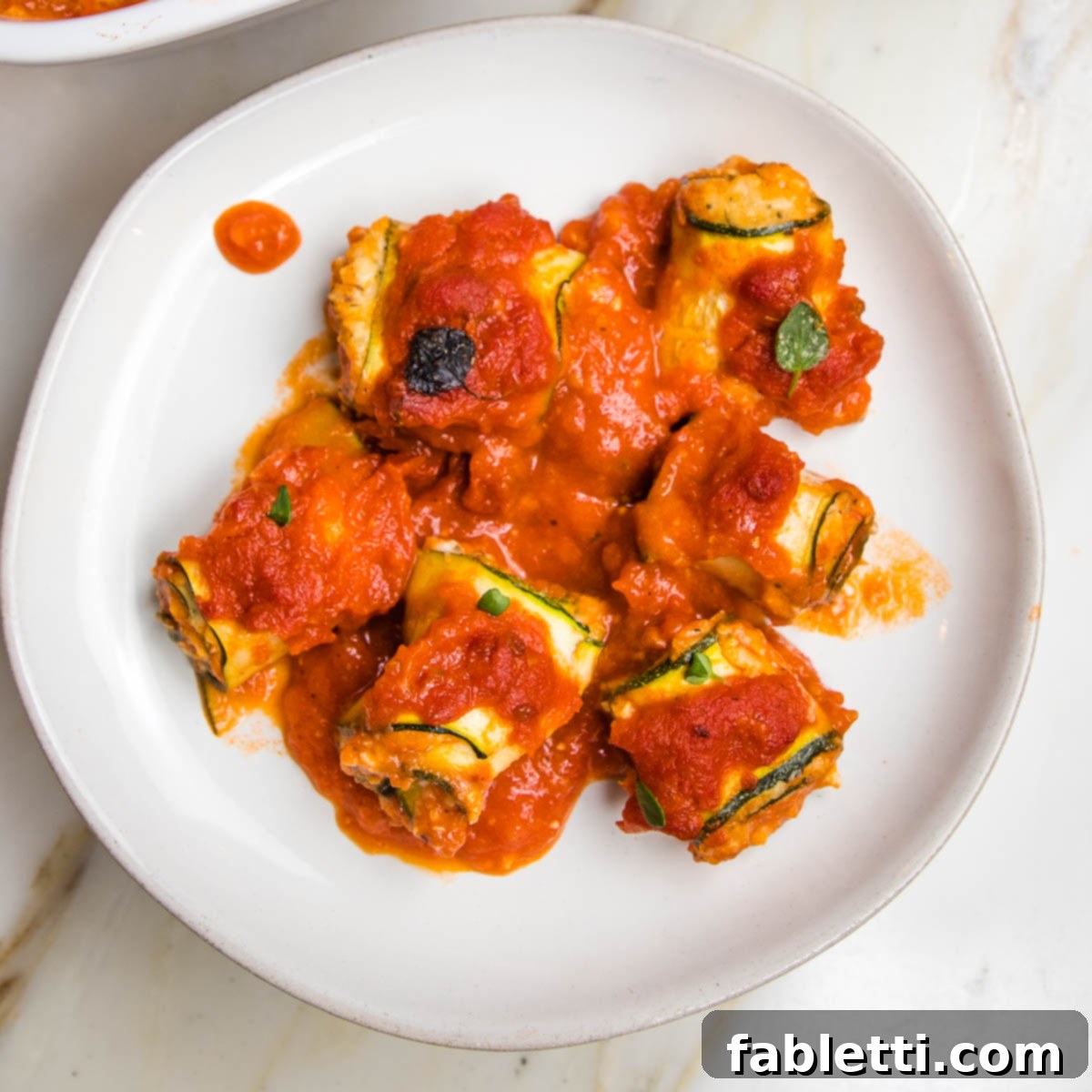 Plant-Based Zucchini Ricotta Rolls 15 White rimmed plate with marinara sauce covered zucchini lasagna roll ups.