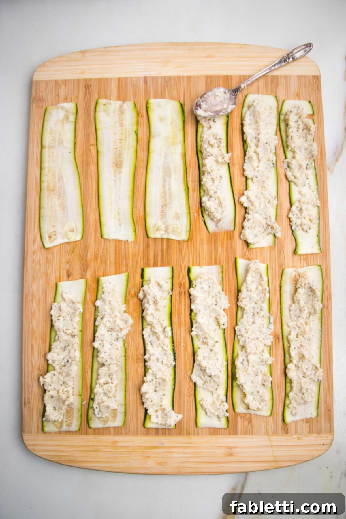 Plant-Based Zucchini Ricotta Rolls 8 Wooden board with zucchini strips laid out, a teaspoon full of ricotta, spread onto each strip is in process.